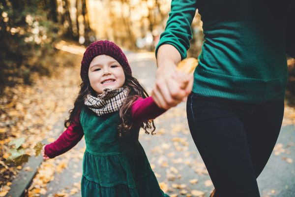 An unrecognizable young mother with a toddler daughter walking in forest in autumn nature, holding hands.