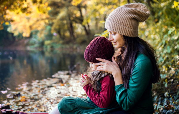 A side view of young mother with a toddler daughter sitting in forest in autumn nature.