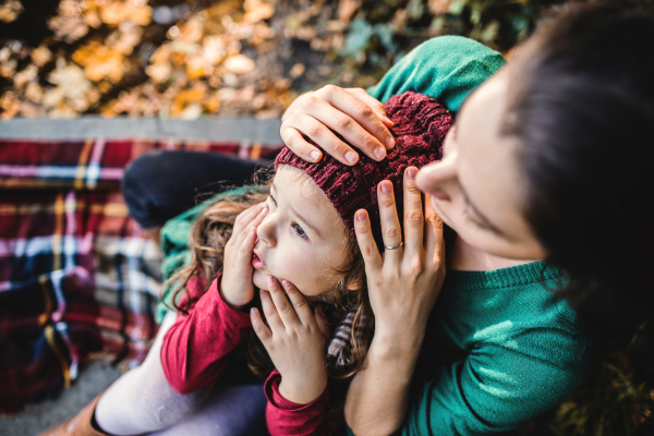 An unrecognizable young mother with a toddler daughter sitting in forest in autumn nature.