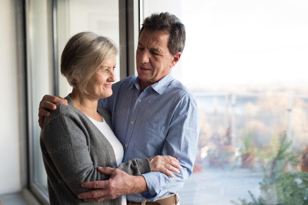 Beautiful senior woman and man in their living room standing at the window, hugging