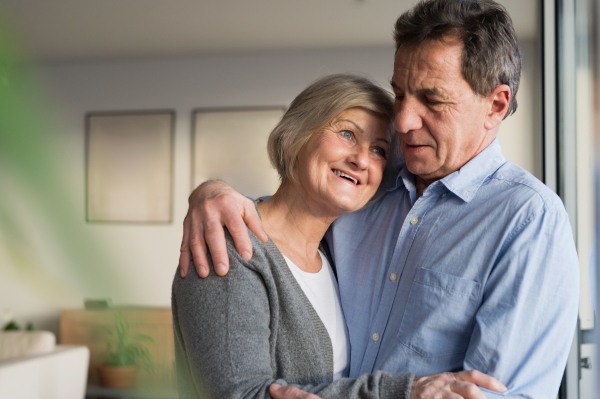 Beautiful senior woman and man in their living room standing at the window, hugging