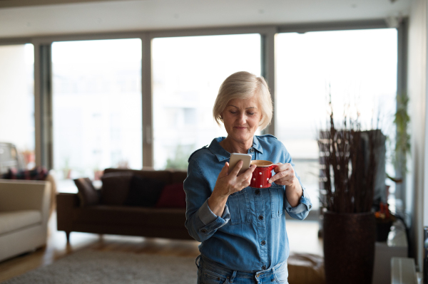 Beautiful senior woman with smart phone and cup of coffee at home in her living room