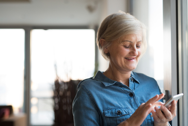 Beautiful senior woman with smart phone in her living room standing at the window