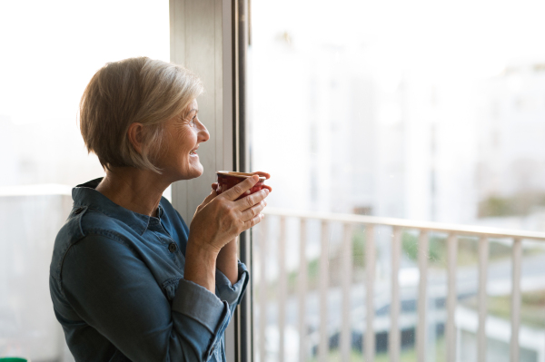 Beautiful senior woman at home standing at the window in her living room holding a cup of coffee or tea, smiling