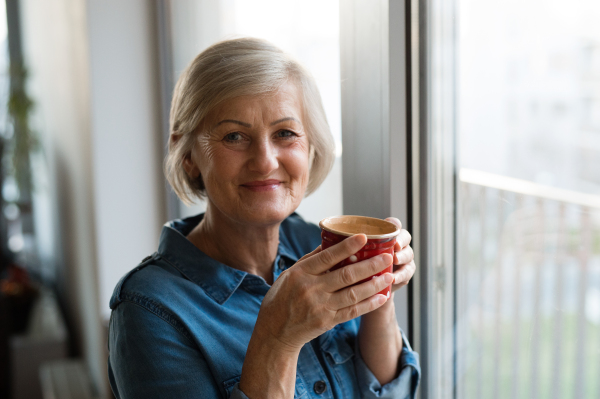 Beautiful senior woman at home standing at the window in her living room holding a cup of coffee or tea, smiling