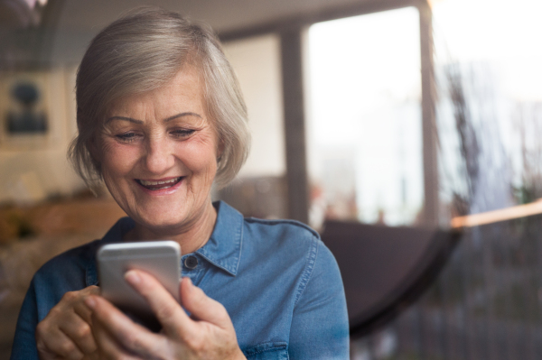 Beautiful senior woman at home in her living room holding smart phone, writing something, view through the window