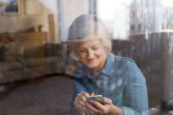 Beautiful senior woman at home in her living room holding smart phone, writing something, view through the window