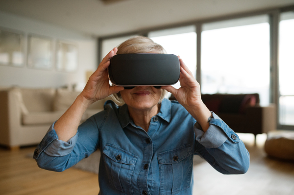 Beautiful senior woman in denim shirt at home in her living room wearing virtual reality goggles