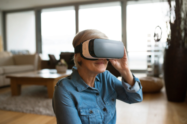 Beautiful senior woman in denim shirt at home in her living room wearing virtual reality goggles