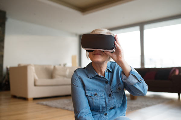 Beautiful senior woman in denim shirt at home in her living room wearing virtual reality goggles