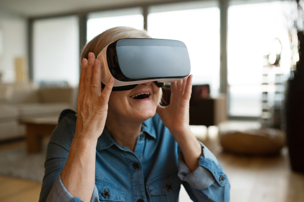 Beautiful senior woman in denim shirt at home in her living room wearing virtual reality goggles