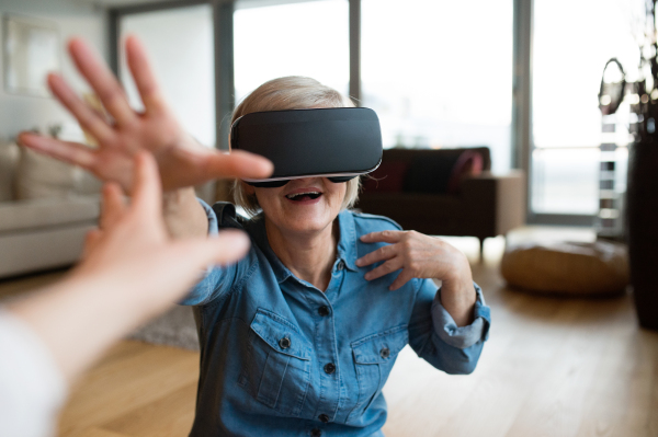 Beautiful senior woman in denim shirt at home in her living room wearing virtual reality goggles, reaching to someone