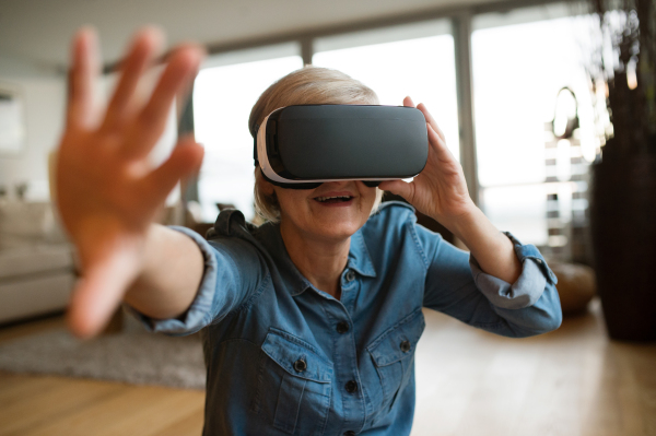 Beautiful senior woman in denim shirt at home in her living room wearing virtual reality goggles, reaching to someone