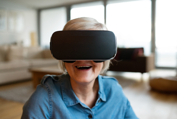 Beautiful senior woman in denim shirt at home in her living room wearing virtual reality goggles