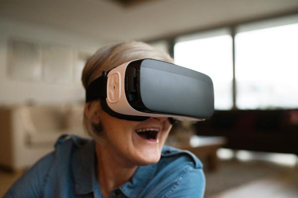 Beautiful senior woman in denim shirt at home in her living room wearing virtual reality goggles