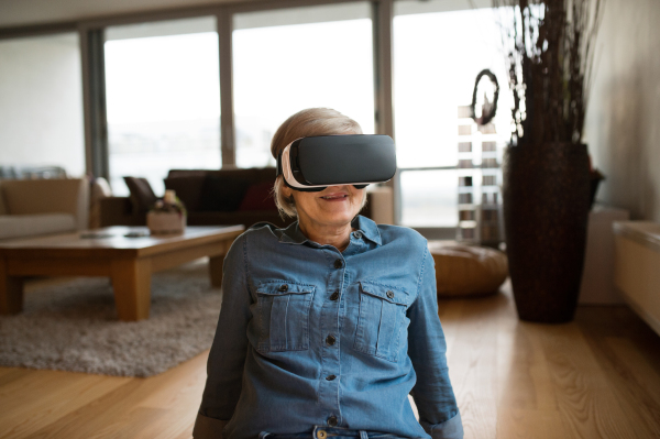 Beautiful senior woman in denim shirt at home in her living room wearing virtual reality goggles