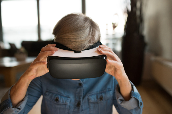 Beautiful senior woman in denim shirt at home in her living room wearing virtual reality goggles, smiling