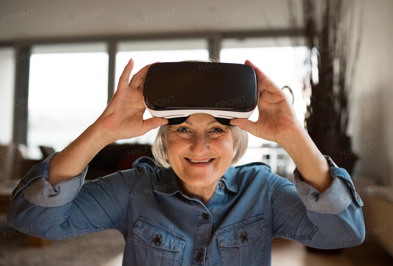 Beautiful senior woman in denim shirt at home in her living room wearing virtual reality goggles, smiling