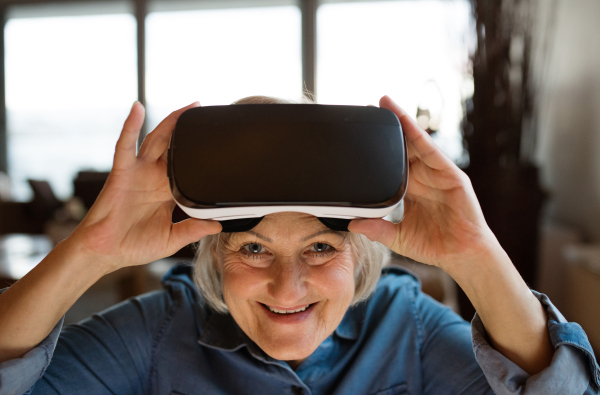 Beautiful senior woman in denim shirt at home in her living room wearing virtual reality goggles, smiling