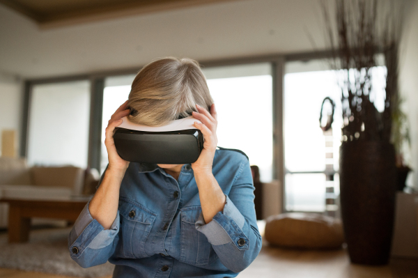 Beautiful senior woman in denim shirt at home in her living room wearing virtual reality goggles