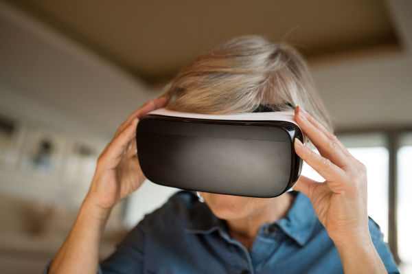 Beautiful senior woman in denim shirt at home in her living room wearing virtual reality goggles