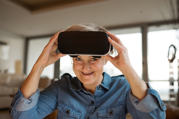 Beautiful senior woman in denim shirt at home in her living room wearing virtual reality goggles