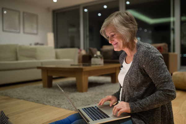 Beautiful senior woman at home in her living room sitting on the floor, working on laptop