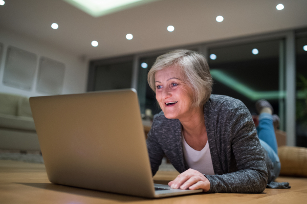 Beautiful senior woman at home in her living room lying on the floor, working on laptop