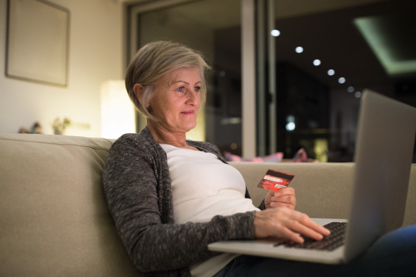 Beautiful senior woman with laptop at home in her living room sitting on the couch, holding a credit card, shopping online