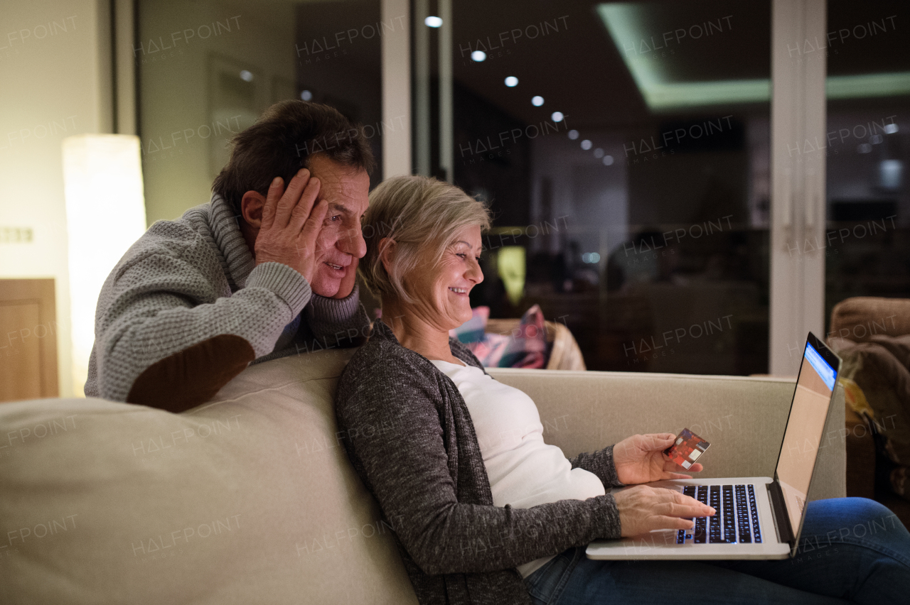 Beautiful senior couple at home in their living room, woman with laptop sitting on couch shopping online