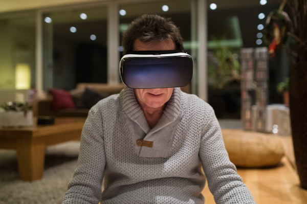 Senior man in gray sweater at home in his living room in the evening wearing virtual reality goggles