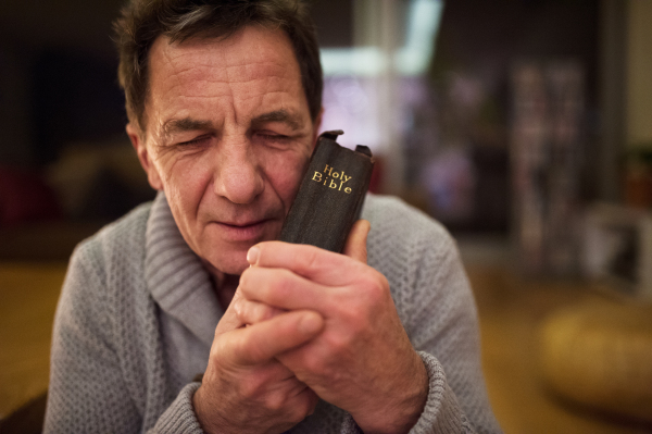 Senior man praying, holding an old Bible in his hands, eyes closed, close up.