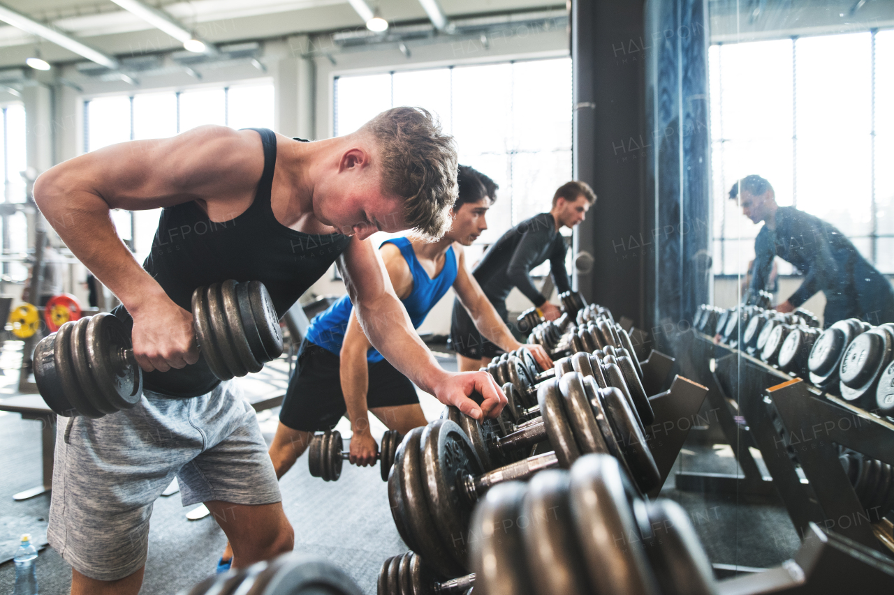 Young fit men doing strength training, exercising with dumbbells in modern gym.