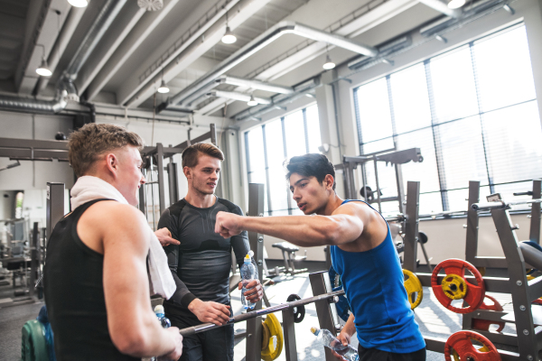 Young friends standing and talking in modern crossfit gym, holding plastic bottles and giving fist bump.