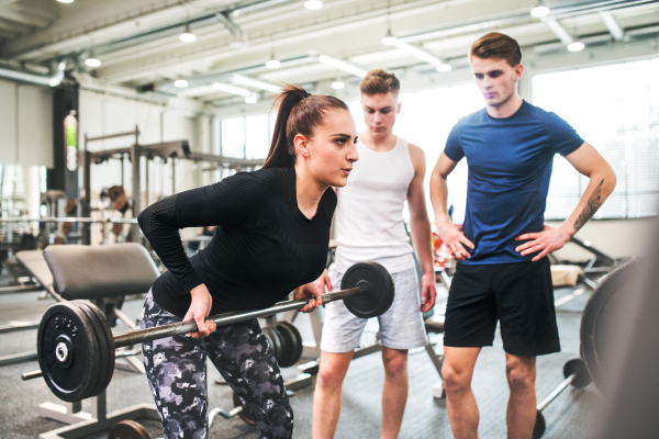 Young handsome fit men in gym looking at their personal trainer working out, lifting bar of barbell, flexing muscles.