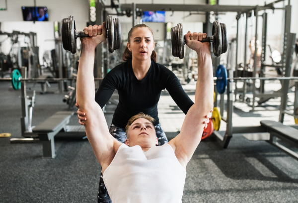 Fit young man in gym with his personal trainer exercising with dumbbells.