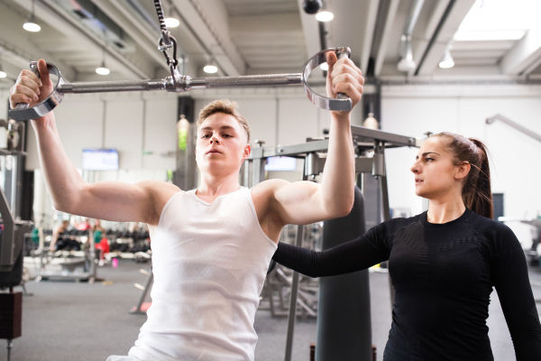 Young handsome fit man with his personal trainer working out on pull-down machine in gym. Bodybuilder exercising with cable weight machine.