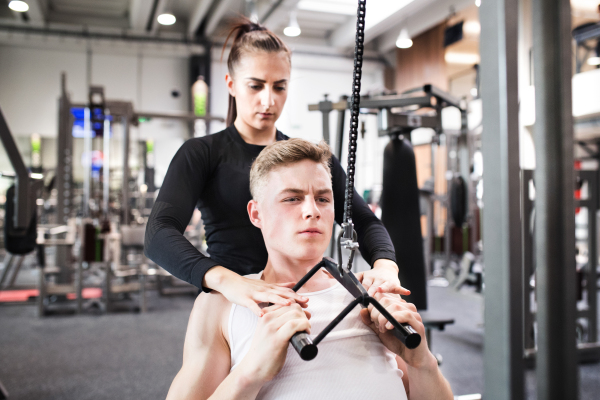 Young handsome fit man with his personal trainer working out on pull-down machine in gym. Bodybuilder exercising with cable weight machine.