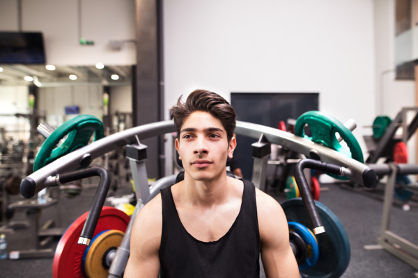 Young fit hispanic man in gym going to work out on fitness machine, flex muscles. Bodybuilder prepared for training, going to do shoulder press.