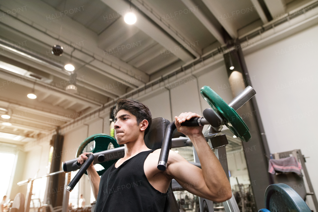 Young fit hispanic man in gym working out on fitness machine, flexing muscles. Bodybuilder training, doing shoulder press.