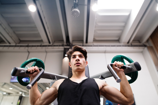 Young fit hispanic man in gym working out on fitness machine, flexing muscles. Bodybuilder training, doing shoulder press.