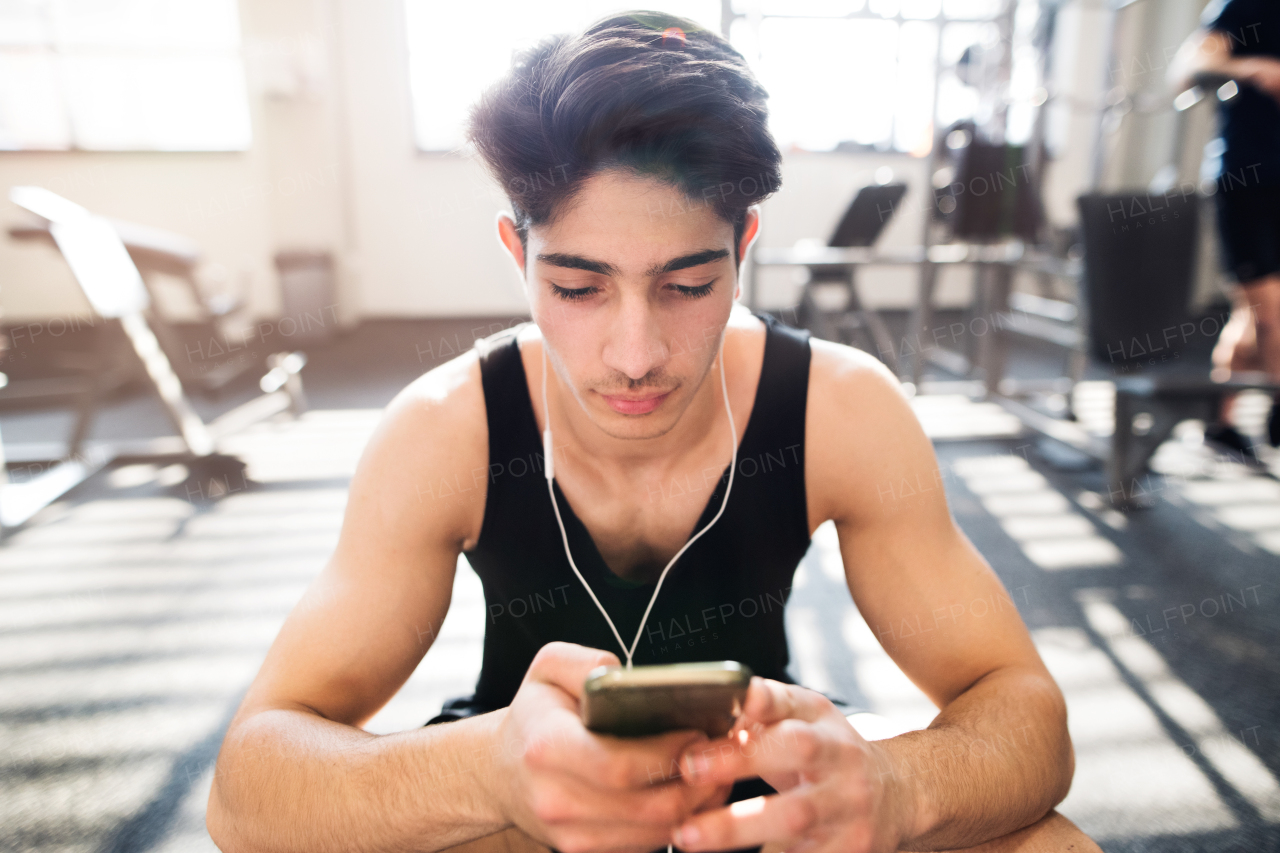 Hispanic fitness man in gym resting, holding smart phone, earphones in his ears listening music