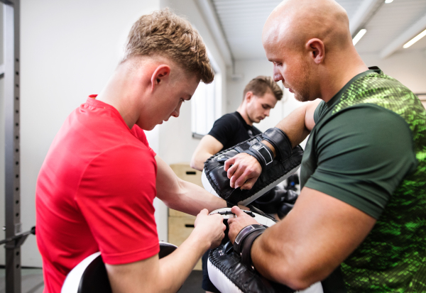 Handsome fit young man in gym with his personal trainer preparing for boxing, putting on boxing gloves.