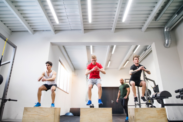 Three young fit men in crossfit gym with their personal trainer doing box squats, practicing box jumps.