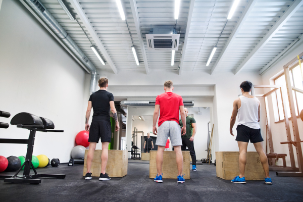 Three young fit men in crossfit gym with their personal trainer doing box squats, practicing box jumps. Rear view.