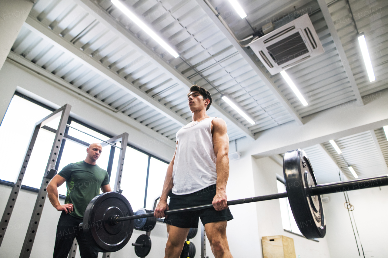 Young handsome fit man with a personal trainer in gym working out, lifting bar of barbell, flexing muscles.