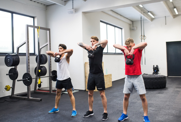 Three young fit men in crossfit gym working out, doing kettlebell swings.