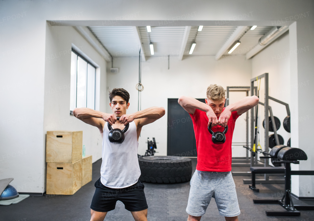 Two young fit men in crossfit gym working out, doing kettlebell swings.