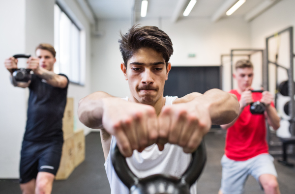 Three young fit men in crossfit gym working out, doing kettlebell swings.