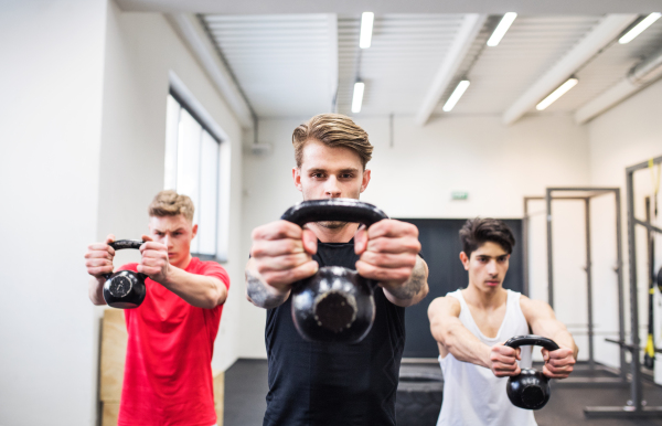 Three young fit men in crossfit gym working out, doing kettlebell swings.